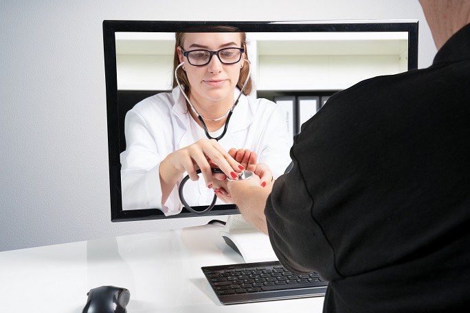 Rückansicht eines Mannes, der vor einem Computerbildschirm sitzt. Der Bildschirm zeigt eine Ärztin, die durch den Bildschirm hindurchgreift und beim Mann den Puls am Handgelenk mit einem Stethoskop misst.