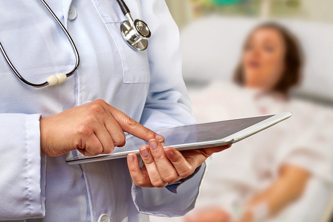 Person im Arztkittel mit Tablet in der Hand im Vordergrund, Frau im Krankenhausbett im Hintergrund