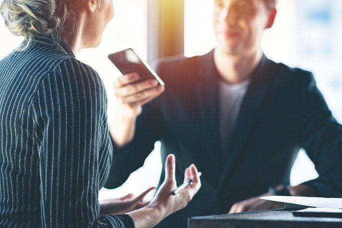 Ein Mann hält einer Frau ein Smartphone wie ein Diktiergerät entgegen. Die Frau ist nur von hinten zu sehen. Sie gestikuliert. Beide tragen Businesskleidung.