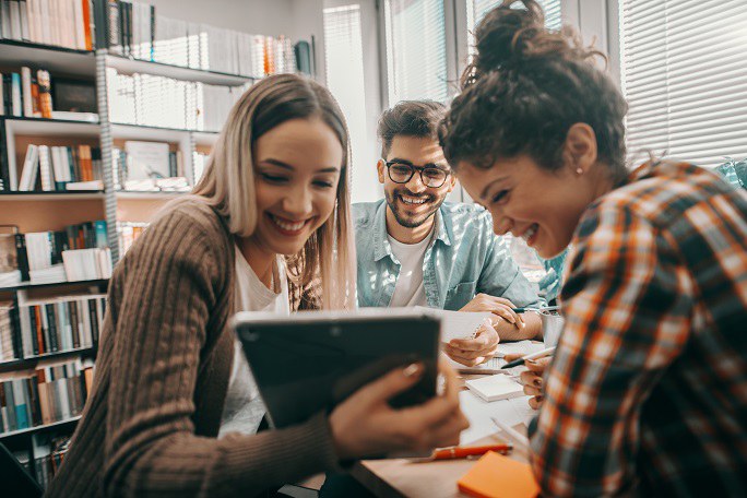 Drei glückliche junge Menschen in Alltagskleidung sitzen an einem Tisch in einer Bibliothek und nutzen gemeinsam ein Tablet für ein Schulprojekt