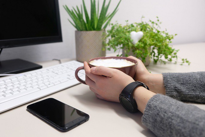 Zwei Hände umschließen ein Heißgetränk. Daneben liegt ein Smartphone. Im Hintergrund befinden sich Pflanzen und ein Computer. 