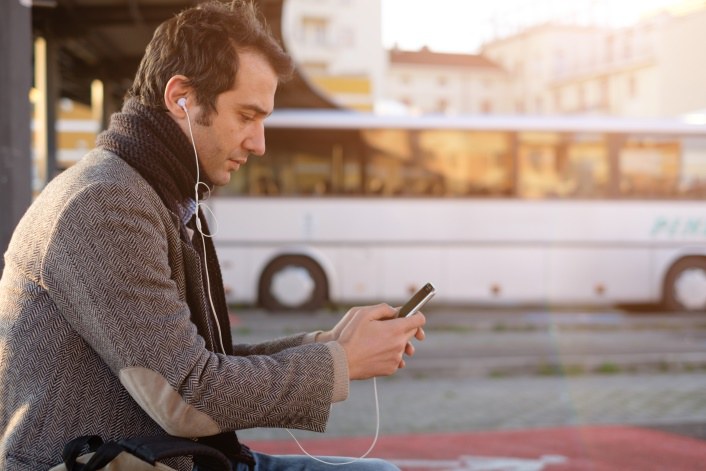 Mann sitzt mit Handy und Kopfhörern an einer Busstation. 