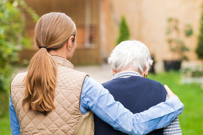 Es sind zwei Personen von hinten abgebildet: Eine jüngere Frau mit langem Haar, die ihren Arm um eine ältere Person mit weißem Haar gelegt hat. Im Hintergrund sind ein Garten und ein Gebäude zu sehen.