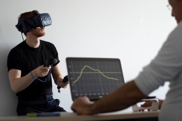 Ein junger Man hat eine VR-Brille an und hält noch zwei kleinere Geräte in beiden Händen. Ein ander Mann schautz sich einen Graphen auf einem Laptop an.