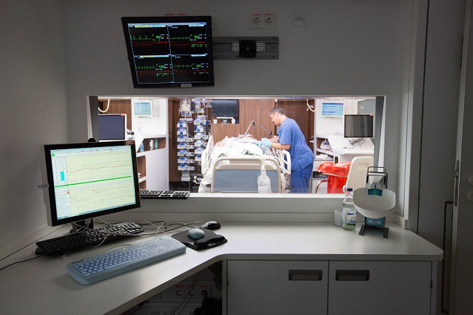 Blick von einem Kontrollraum in ein Zimmer auf der Intensivstation, in dem sich ein Patient und ein Pfleger befinden.