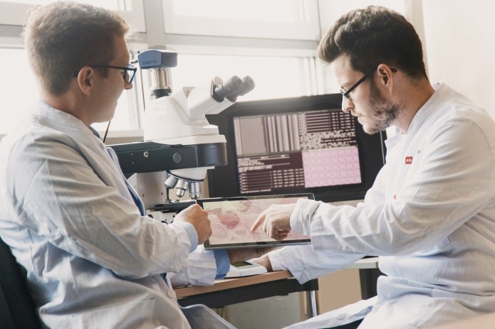 Zwei Männer schauen sich Gewebeproben auf einem Tablet an. Im Hintergrund steht ein größerer Bildschirm sowie ein Mikroskop.