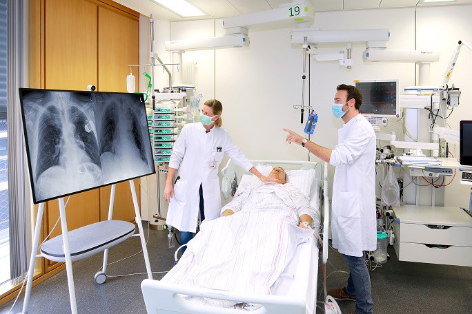 Ärztin und Arzt stehen am Bett eines Patienten und sehen sich Röntgenbilder des Thorax an.