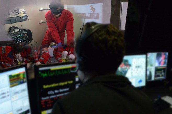 Eine Person sitzt mit Headset vor mehren Bildschirmen, die unterschiedliche Daten anzeigen. Diese Person schaut durch ein Fenster in einen separaten Raum, wo eine Person eine VR-Brille trägt und an Trainingspuppen übt. 