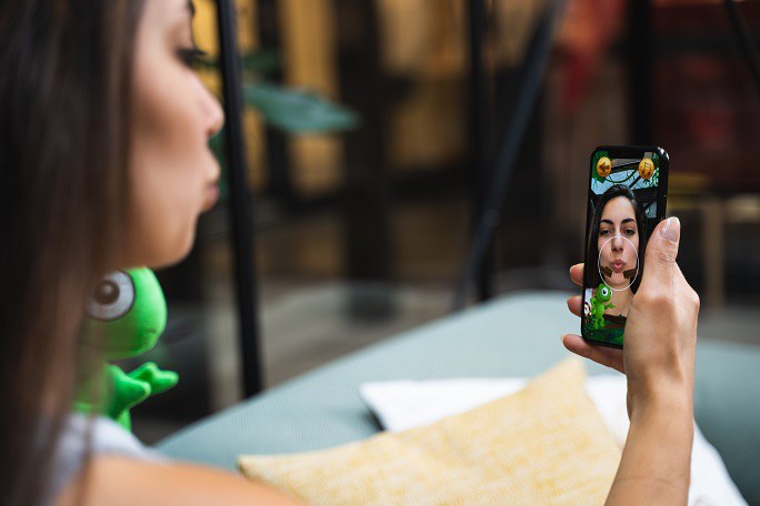 Eine Frau filmt sich selbst mit einem Smartphone. Auf dem Bildschirm ist ein kleines grünes Wesen und ein Kreis um den Mund der Nutzerin eingeblendet.