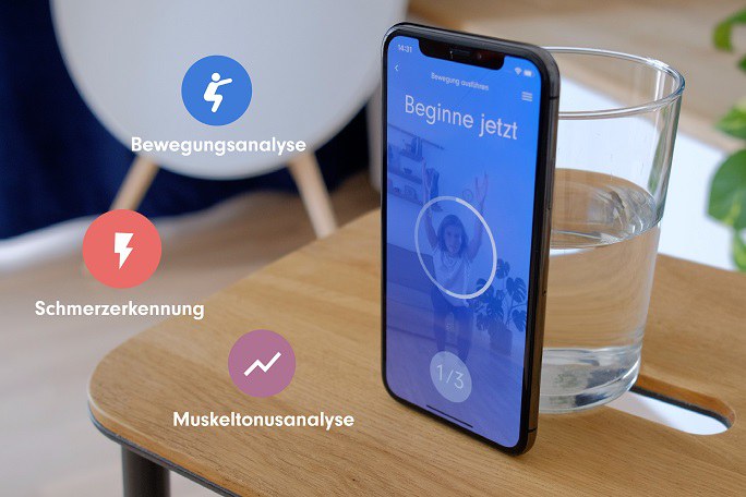 Ein Smartphone steht an ein Wasserglas geleht auf einem Stuhl. Auf dem Bildschirm ist ein Foto einer Frau zu sehen, die eine Sportübung macht und darüber steht der Text "Beginne jetzt". Neben dem Smartphone sind drei Symbole eingeblendet, die mit den Worten "Bewegungsanalyse", "Schmerzerkennung" und "Muskeltonusanalyse" beschriftet sind.