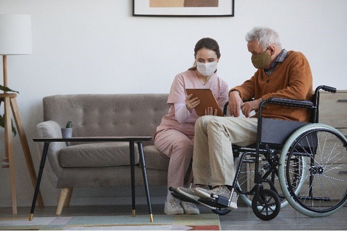 Pflegerin und Ältere Person im Rollstuhl schauen gemeinsam auf ein Tablet.