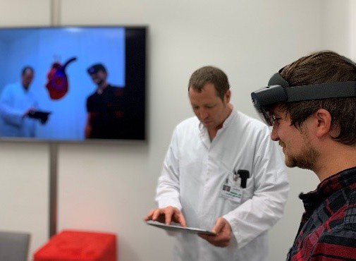 Ein Arzt mit Tablet und ein Patient mit VR-Brille. Im Hintergrund sieht man die beiden mit dem Modell eines Herzen auf einem Bildschirm.