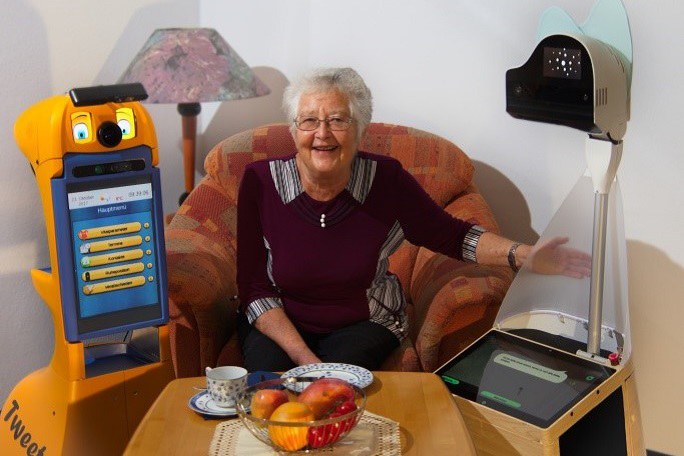 Eine ältere Dame sitzt in einem Wohnzimmersessel. Rechts von ihr steht ein abstrakter Roboter und links von ihr ein Roboter mit Gesicht und großem Bildschirm.