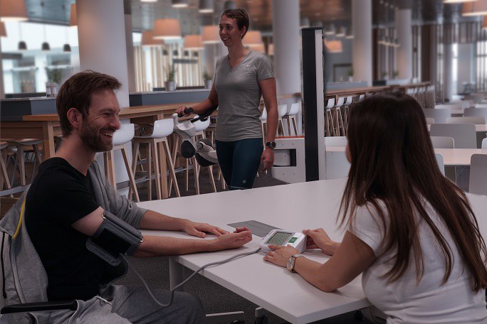 Im Vordergrund sitzen eine junge Frau und ein junger Mann an einem Tisch. Sie misst seinen Blutdruck. Im Hintergrund läuft eine weitere Frau. Ihr rechter Arm wird von einem Roboter gestützt, der sie beim Gehen begleitet.