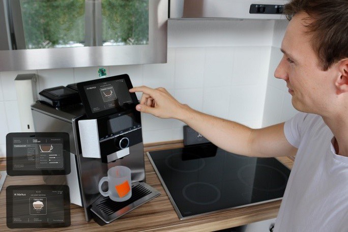 Ein junger Mann interagiert mit einer Kaffemaschine mit einem komplexen Display, die in einer Küche steht.