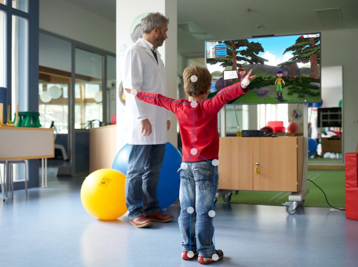Ein Kind ahmt die Bewegungen nach, die er auf einem Bildschirm sieht. Neben ihm steht ein Arzt und schaut zu.