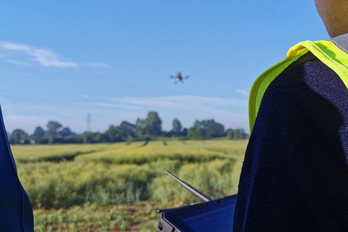 Mann steht mit Steuerung auf einem Feld und lenkt eine Drohne am Himmel.