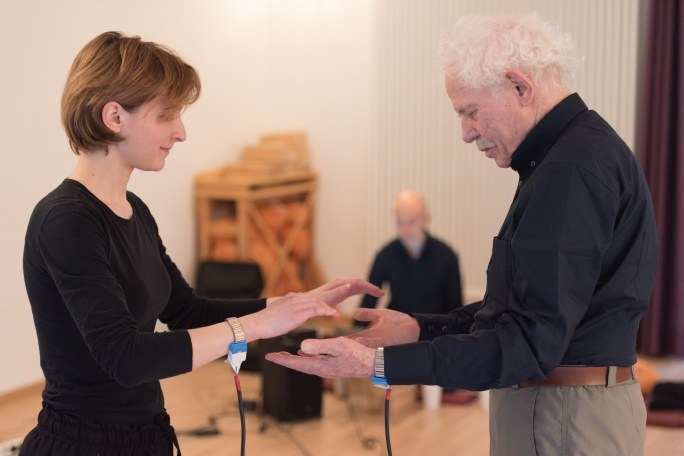 Eine Frau hält ihre ausgestreckten Hände über die Hände eines älteren Mannes, der ihr gegenüber steht. Beide haben ein verkabeltes Armband am Handgelenk.