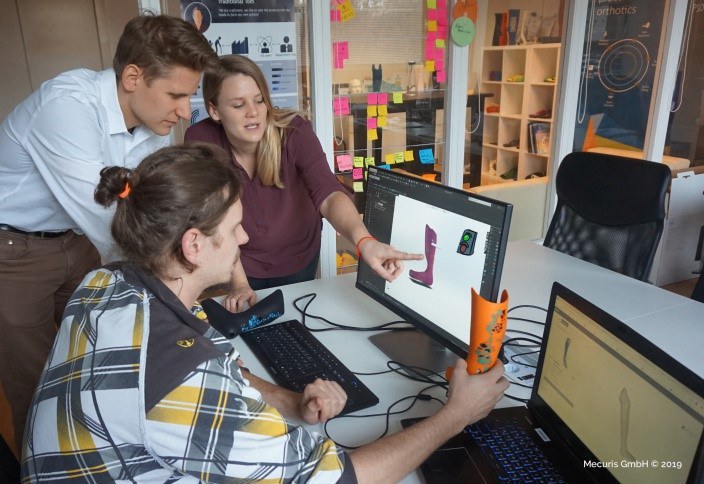 Drei junge Menschen sehen sich die Abbildung einer Orthese auf einem Computerbildschirm an. Einer der drei hält ein verkabeltes, echtes Modell der Orthese in der Hand.