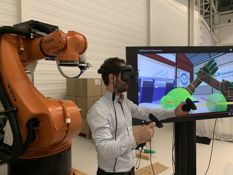 Ein Mann hat eine VR/AR-Brille an und hält zwei verkabelte Gegenstände in der Hand. Hinter ihm steht eine große Maschine und neben ihm ein Bildschirm, wo diese Maschine zu sehen ist.