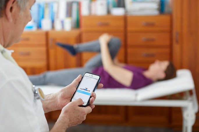 Ein Arzt hält ein Smartphone in der Hand, auf der eine Übung angezeigt wird. Im Hintergrund befindet sich eine Patientin, die diese Übung auf einer Liege ausführt.