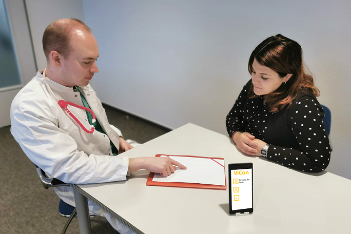Ein Arzt sitzt einer Frau an einem Tisch gegenüber und deutet euch eine vor ihm liegende Mappe im A4-Format und ein Smartphone, welches ViCon anzeigt.
