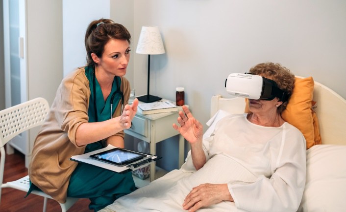 Eine ältere Dame liegt im Bett und trägt eine VR-Brille. Eine Ärztin sitzt mit einem Tablet neben ihr. Beide bewegen eine Hand zueinander.