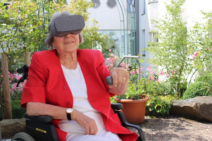 Eine ältere Frau sitzt mit einer VR-Brille auf einem Stuhl. Außerdem hält Sie in der Hand eine kleine Fernbedienung.