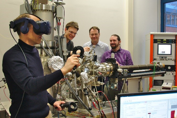 Ein Mann mit VR-Brille und Steuerelementen in beiden Händen steht in einem Labor. Im Hintergrund sind drei Männer zu sehen, die seine Handlungen beobachten.