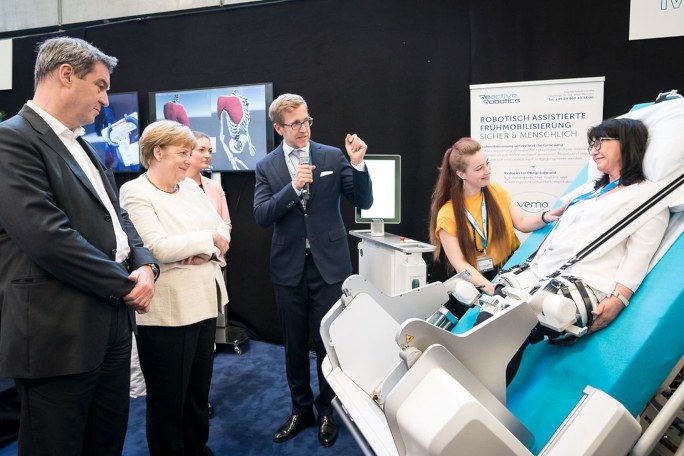 Bundeskanzlerin Angela Merkel und der Bayerische Ministerpräsident Markus Söder stehen vor Frau, die die adaptive Robotik am Krankenbett austestet